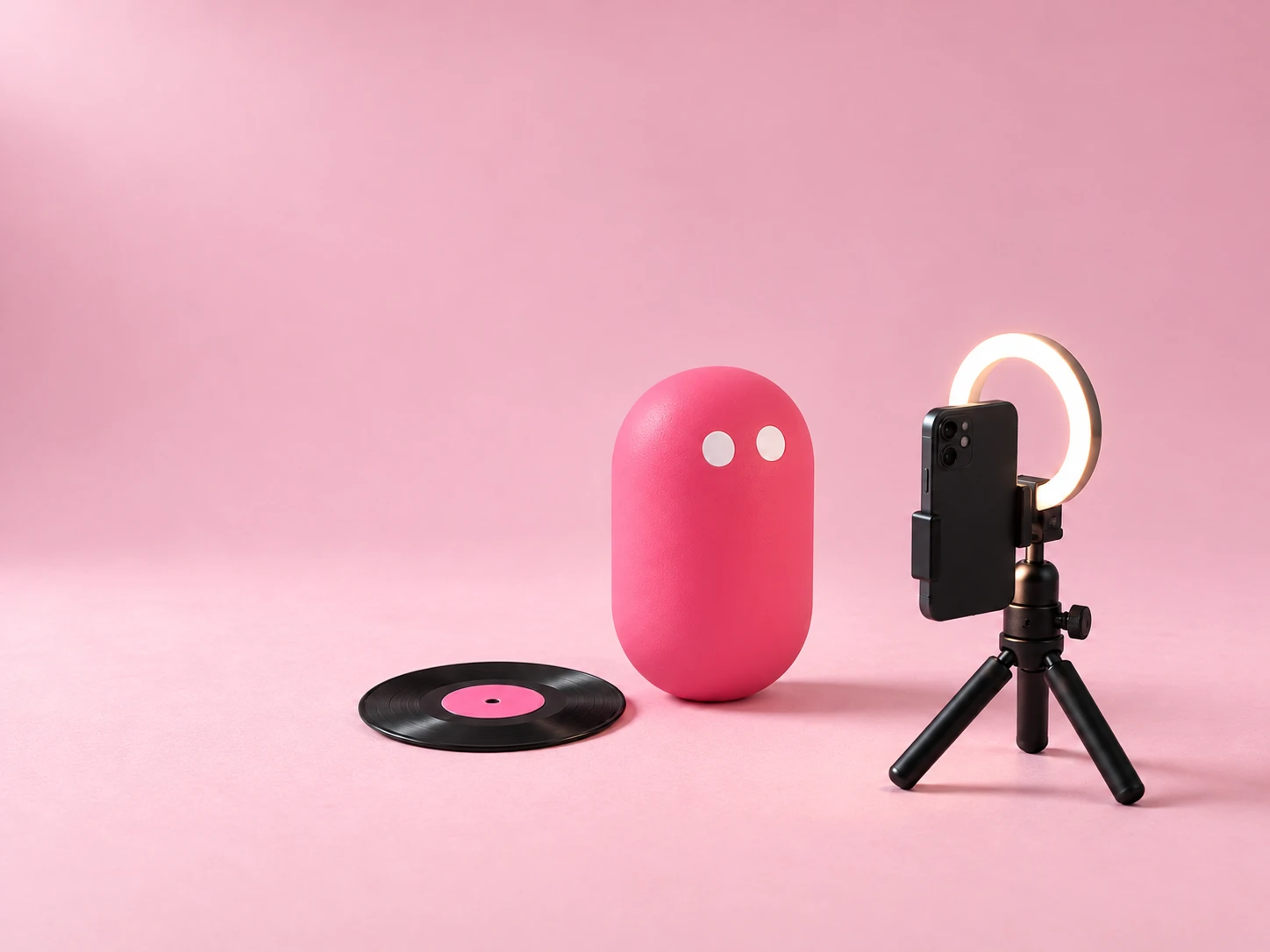 A wide-eyed pink pill in front of a phone on a tiny tripod, with a ring light and a small vinyl record.